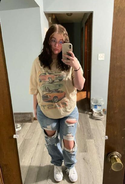 Emily taking a mirror selfie wearing a graphic tee, ripped jeans, and white sneakers indoors.