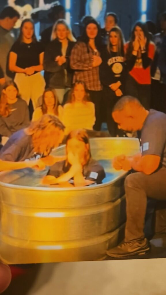 Emily being baptized in a metal tub, surrounded by people watching and two others assisting with the ceremony.
