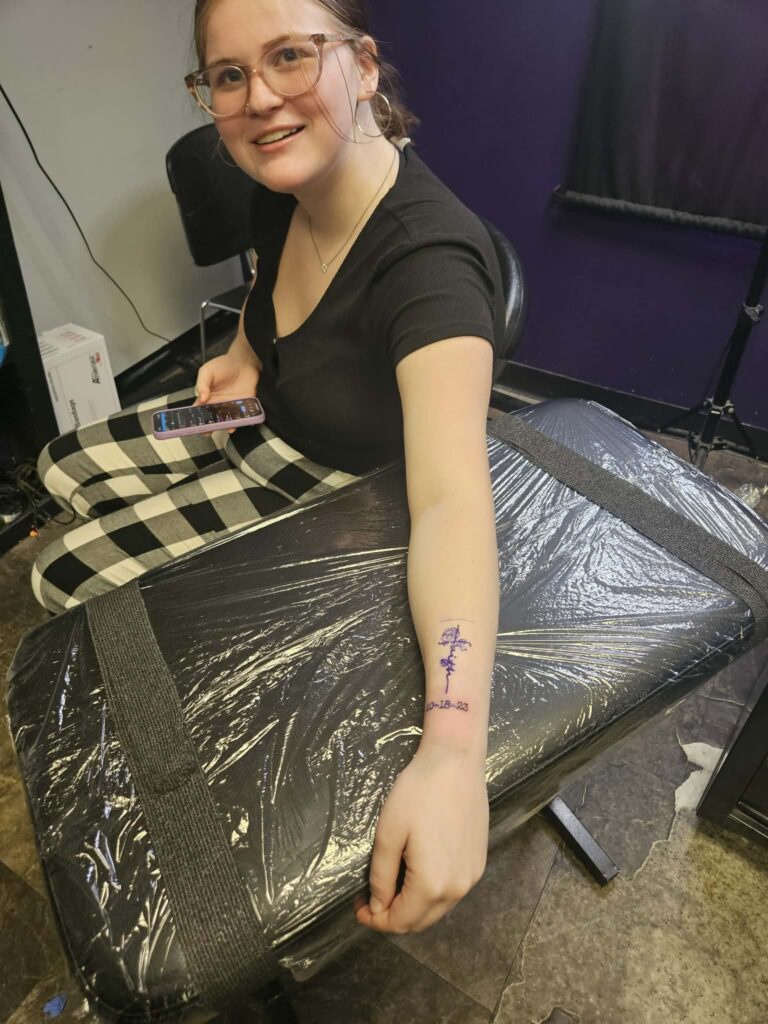 Emily shows her forearm with a fresh purple ink tattoo outline and a date.