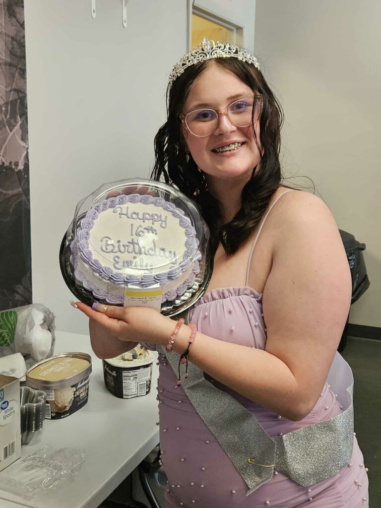 Emily wearing a tiara and sash smiles while holding a birthday cake that reads "Happy 16th Birthday Emily."