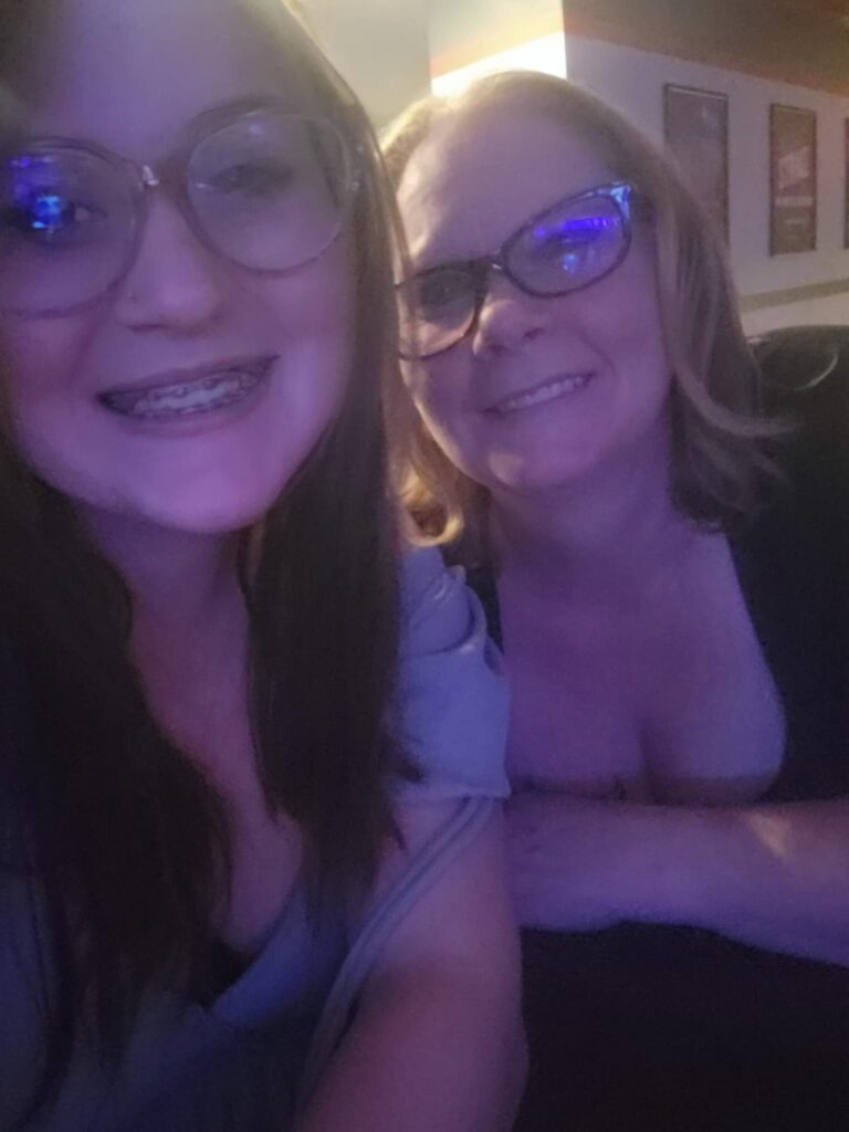 Emily and her mom wearing glasses, taking a close-up smiling selfie in a dimly lit indoor setting.