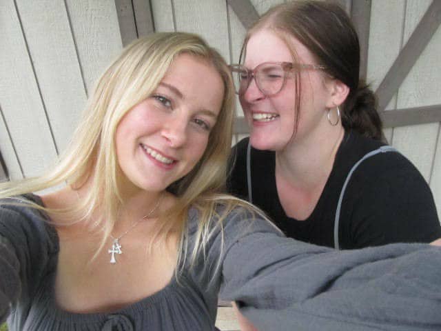 Emily and her sister smiling, taking a selfie, standing in front of a wooden wall with cross beams.