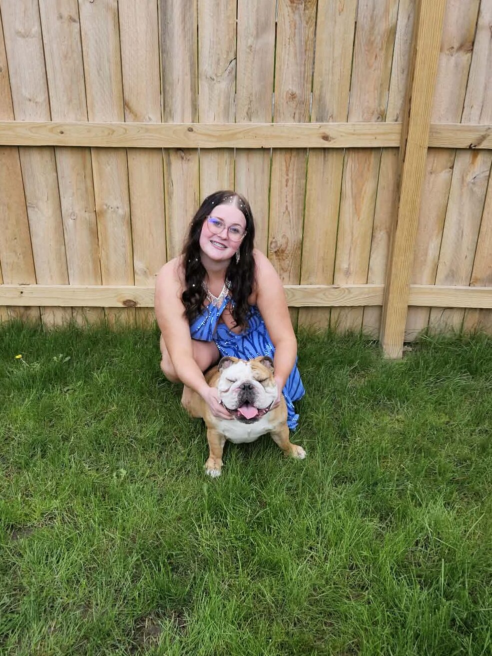 Emily in blue dress crouching on grass, smiling and holding a happy bulldog in front of a wooden fence.