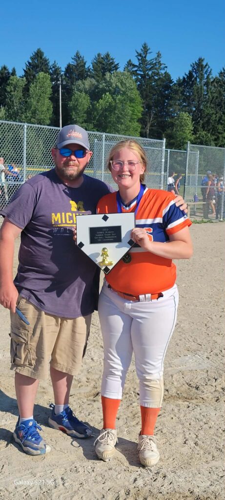 Emily in orange uniform holding a plaque, standing next to a man on a sunny field with a chain-link fence background.