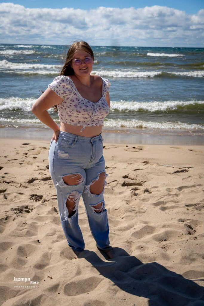 Emily wearing glasses, a floral crop top, and ripped jeans, standing barefoot on a sandy beach with ocean waves behind.